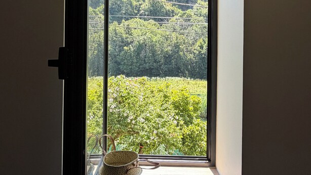 Quarto com janela que dá para uma área arborizada, uma cesta de palha no parapeito e uma porta entreaberta.
