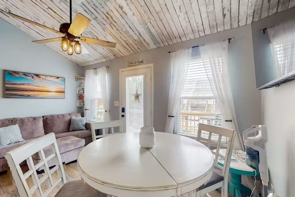 A room with a round table, a ceiling fan, a sofa, and a window with curtains.