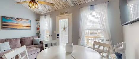 A room with a round table, a ceiling fan, a sofa, and a window with curtains.