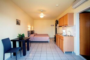 Quarto compacto com cama, uma mesinha com cadeira, cozinha compacta e ventilador de teto.