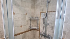 Une douche fermée par des parois en verre, dotée d’un pommeau de douche à main, d’un robinet mural et d’une petite étagère.