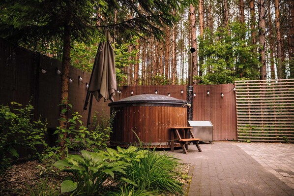A wooden hot tub with a bench, surrounded by greenery and a wooden fence.