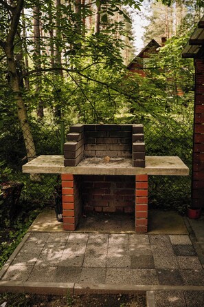 A brick-built outdoor grill with a flat top, surrounded by a chain-link fence, set in a wooded area with a paved walkway.
