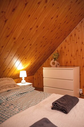 A bedroom with a sloped ceiling, a bed with patterned bedding, a dresser, and a bedside lamp.