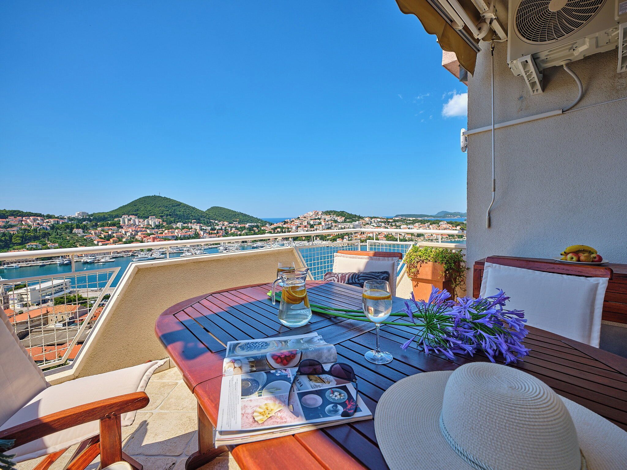 Un balcón con una mesa para dos, con vista a un puerto deportivo y al paisaje urbano.