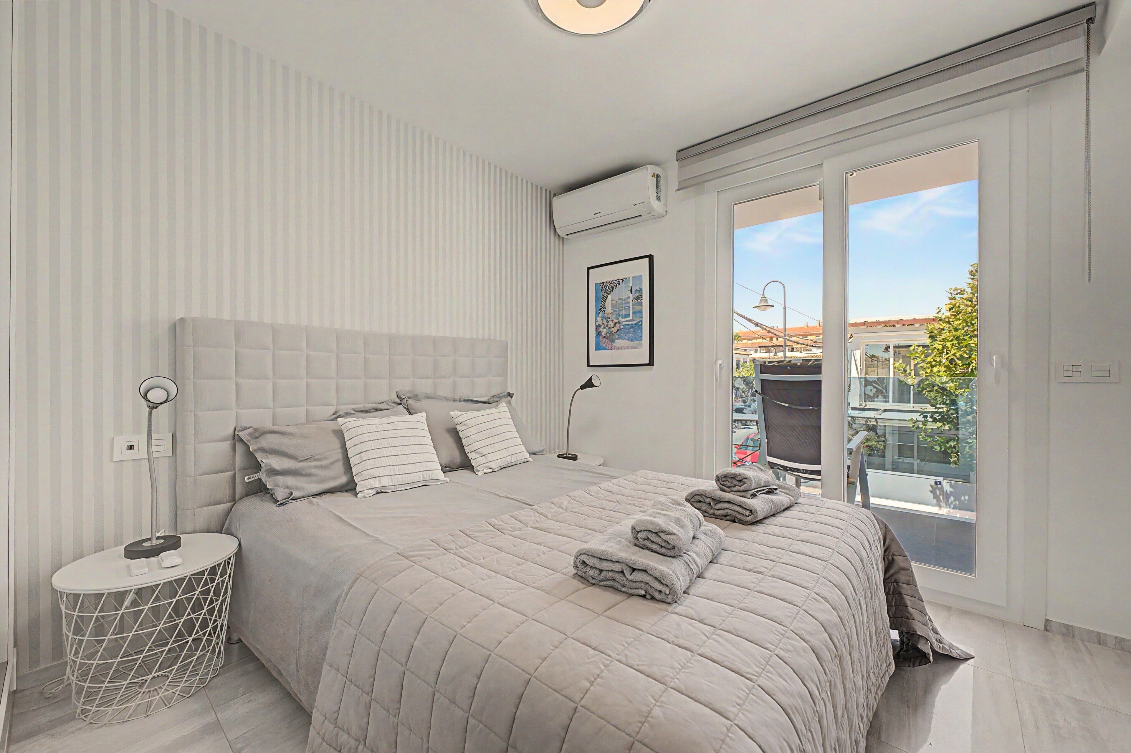 A modern bedroom with a large bed, a bedside table, a wall-mounted air conditioner, and a window with a view of the city.