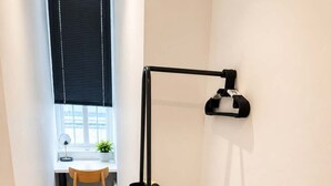 A narrow hallway with a wooden floor, a desk with a chair, a window with blinds, and a black metal rack with a weight plate. - Urban Lux Apartment | Heart of Town (Warrington)