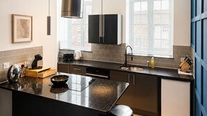 A modern kitchen with a central island, black countertops, a stainless steel range hood, and a large window with blinds. - Urban Lux Apartment | Heart of Town (Warrington)