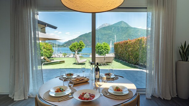 A dining table set with plates, glasses, and a bottle, overlooking a scenic outdoor area with a mountain and water. - Lakefront villa with a private garden and a 4-car garage (Calozzo)