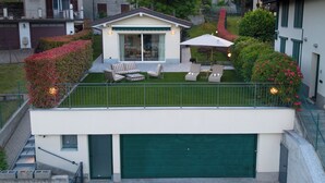 A modern two-story house with a flat roof, a covered patio with outdoor furniture, and a green garage door. - Lakefront villa with a private garden and a 4-car garage (Calozzo)