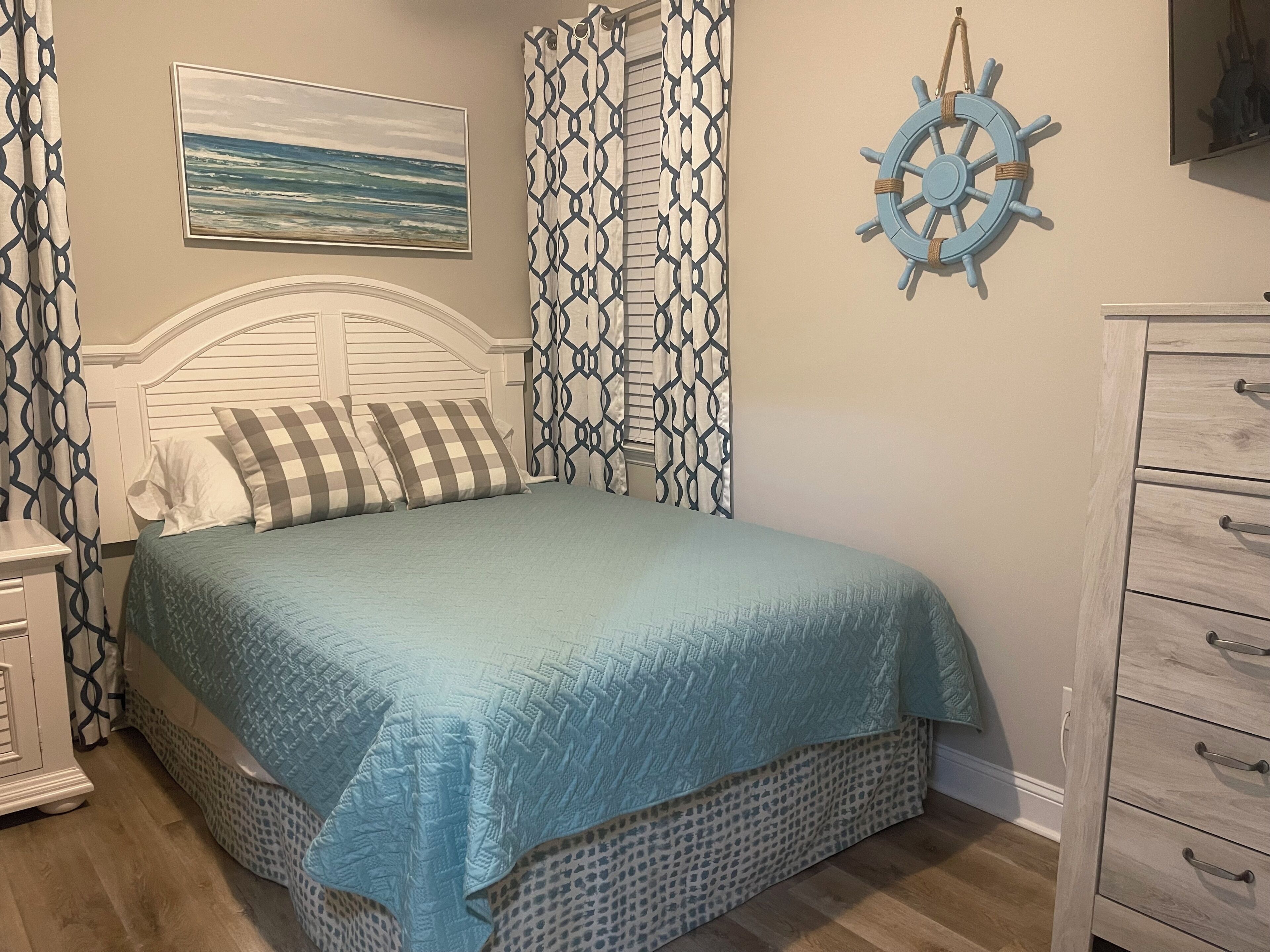 Un dormitorio con una cama, un aparador, un timón anclado a la pared y un cuadro del océano.