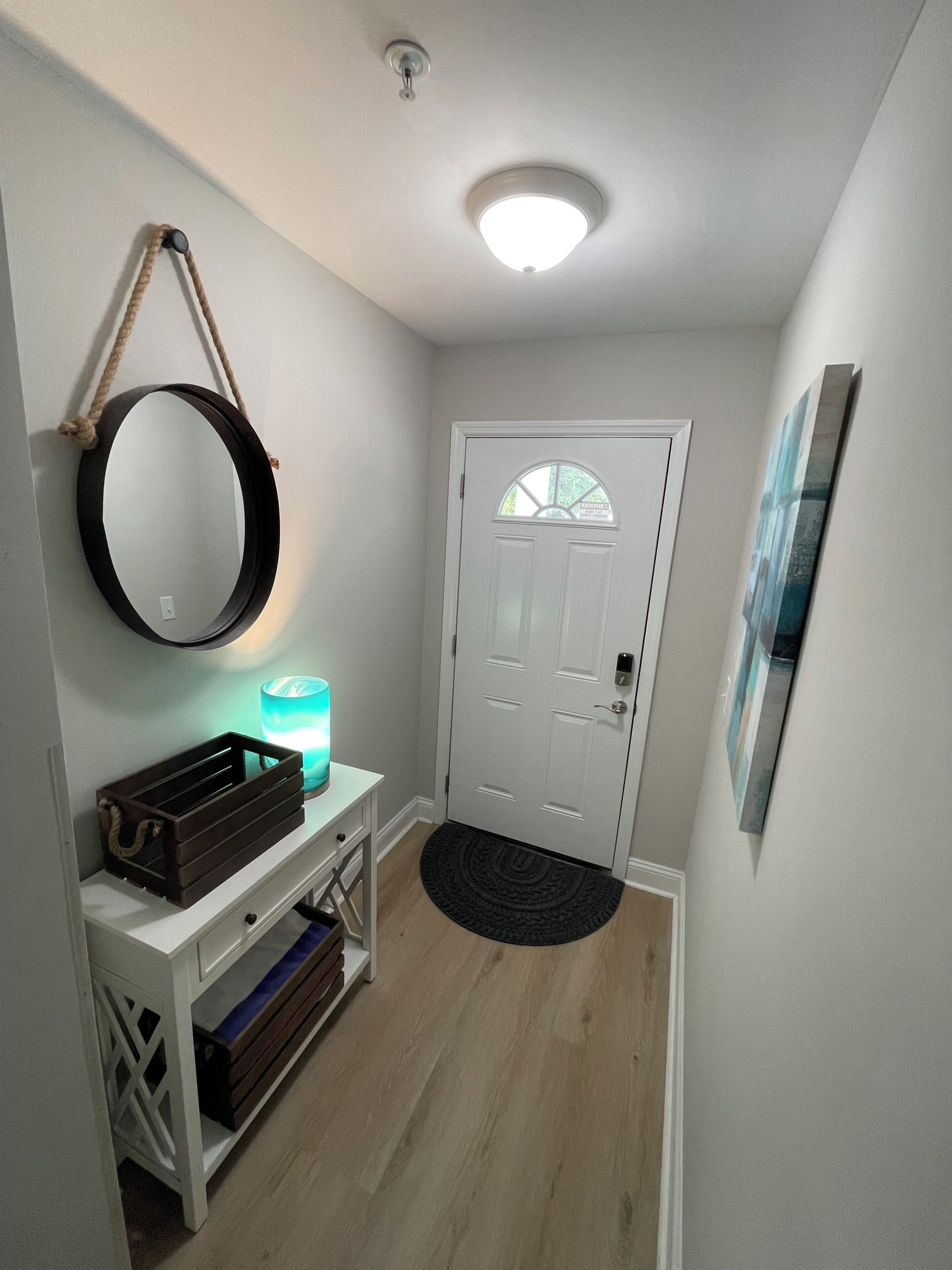 A hallway with a white door, a round mirror, a small table with a candle, and a black rug.