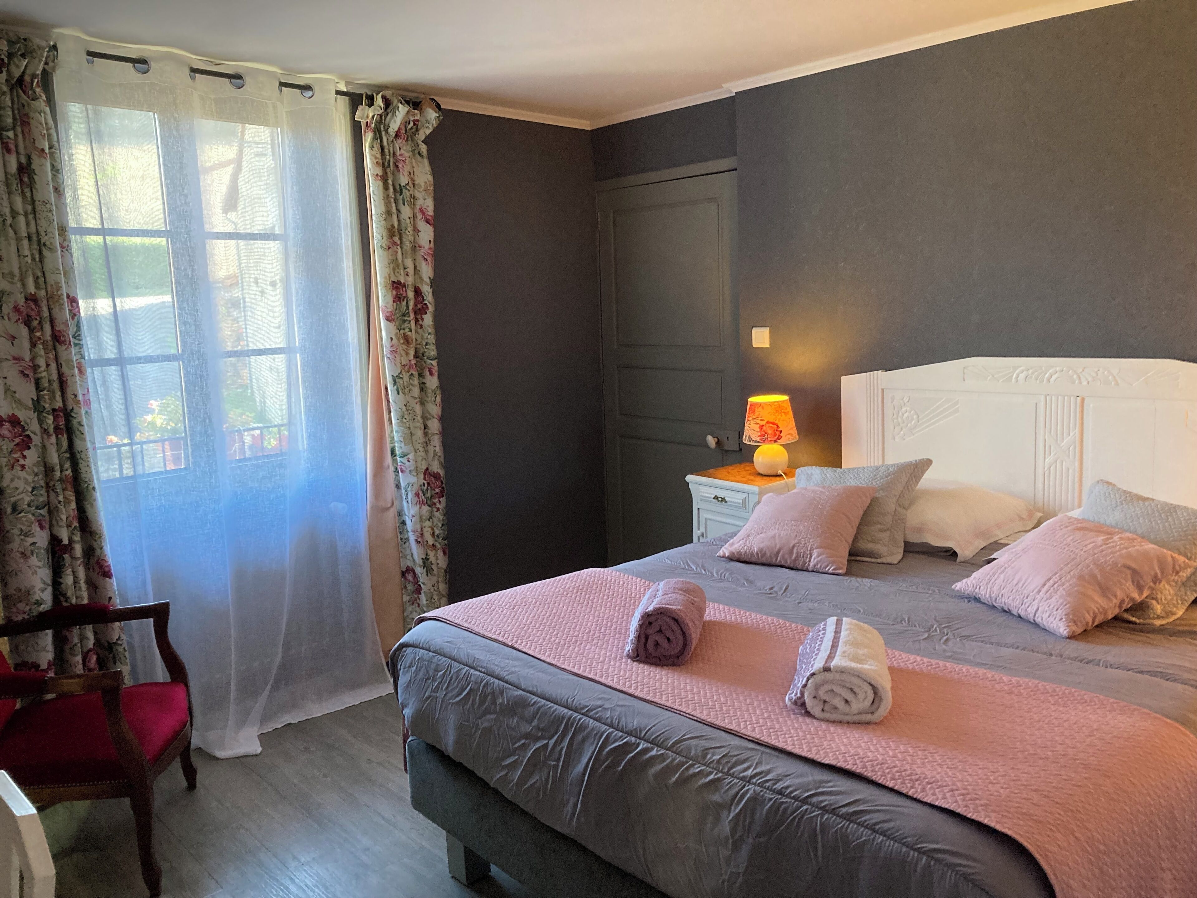 A bedroom with a bed, pillows, a bedside table with a lamp, a chair, and a window with curtains.