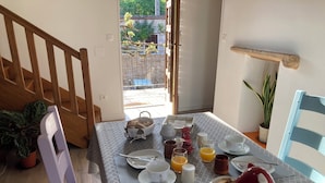Una mesa de desayuno con platos, tazas y una tetera roja, una silla de madera y una planta en maceta.