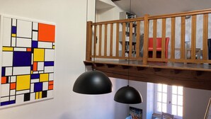 Una habitación con un cuadro abstracto lleno de color, una mesa de comedor con sillas, una barandilla de madera y una ventana con luz natural.