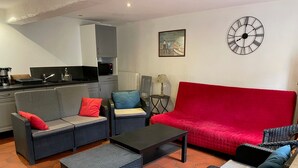 Un área de estar con un sofá rojo, dos sofás grises, una mesa de centro, sillas de mimbre, un reloj y una cocina al fondo.