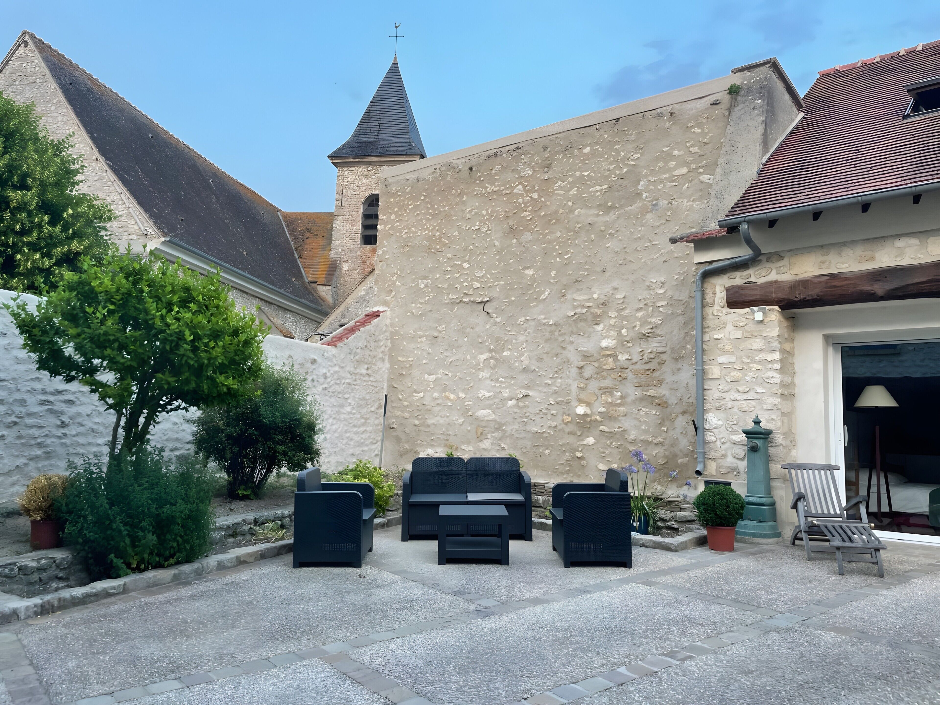 Une cour avec des sièges en plein air, un bâtiment en pierre doté d’une tour et un ciel dégagé.