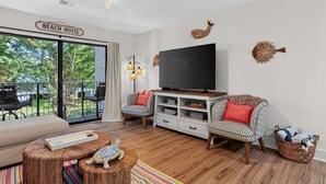 Een woonkamer met een televisie, gestreepte stoelen, een houten salontafel en een bordje met de tekst 'BEACH HOUSE'.