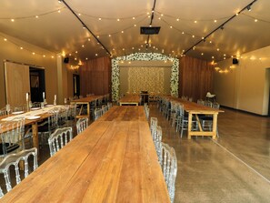 Een ruime zaal met lange houten tafels en transparante stoelen, sfeerverlichting met lichtkoorden en een bloemenachtergrond.