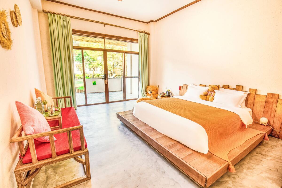 A bedroom with a large bed, a wooden chair, a window with curtains, and a view of greenery outside.