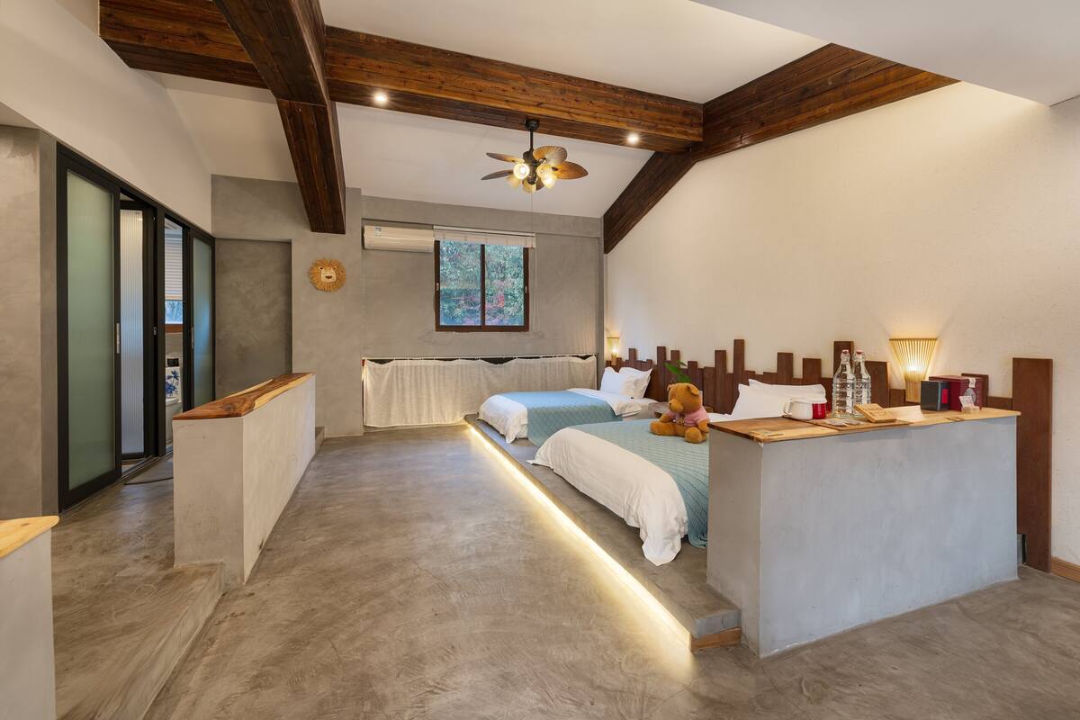 A bedroom with two beds, a wooden ceiling, a bedside table with a lamp, and a window with a view.
