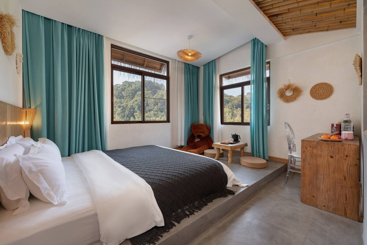 A bedroom with a bed, a chair, a small table, a wooden cabinet, and a window with a view of trees.