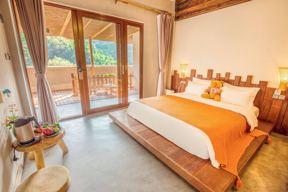 A bedroom with a bed, a small table with a kettle, and a view of the outdoors.