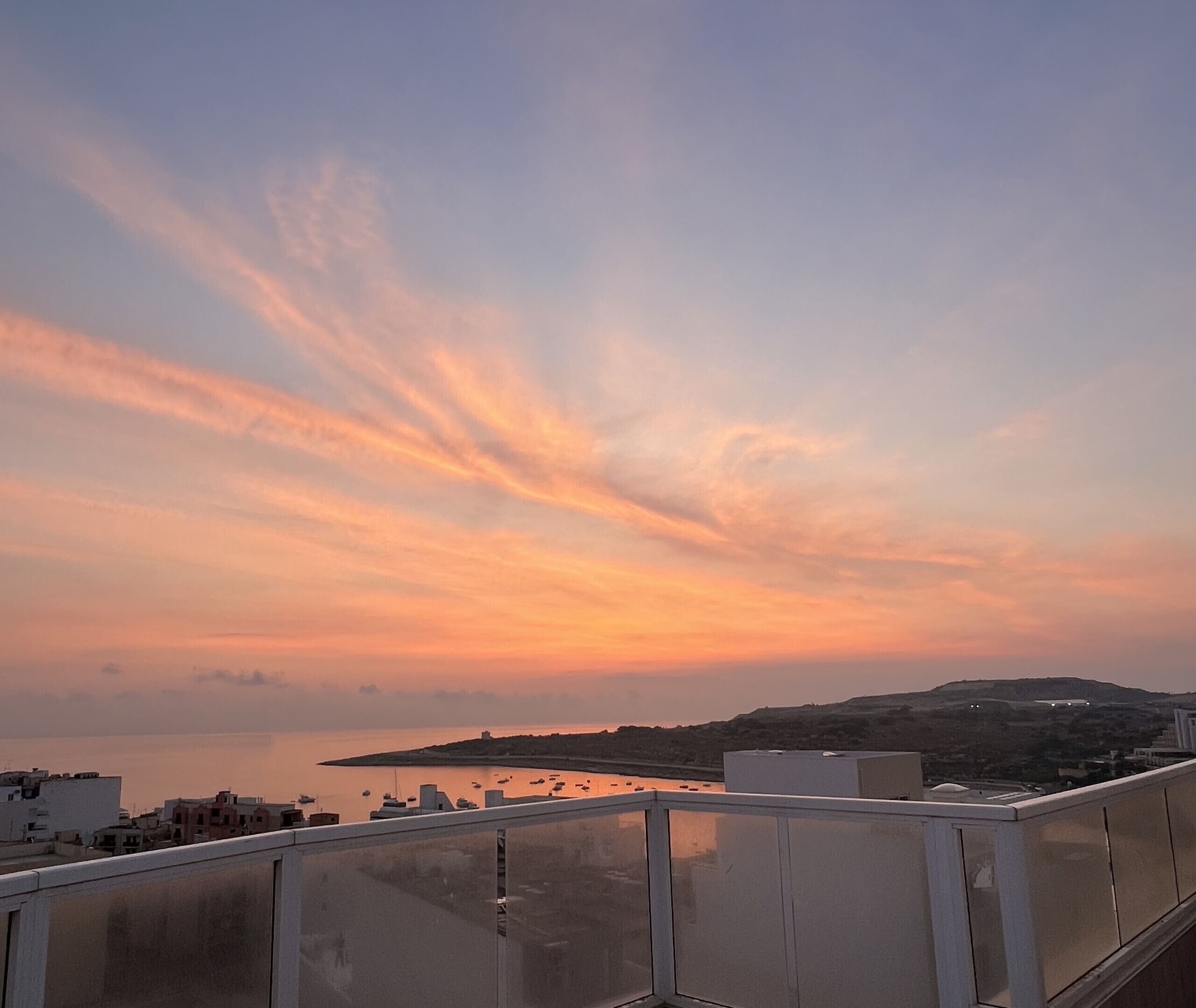 Sonnenuntergangblick von einem Balkon mit Geländer, mit Blick auf ein ruhiges Meer und ferne Hügel.
