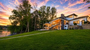 A modern two-story house with a large front lawn, a stone chimney, and a view of a lake at sunset. - Willow Landing (Troutman)