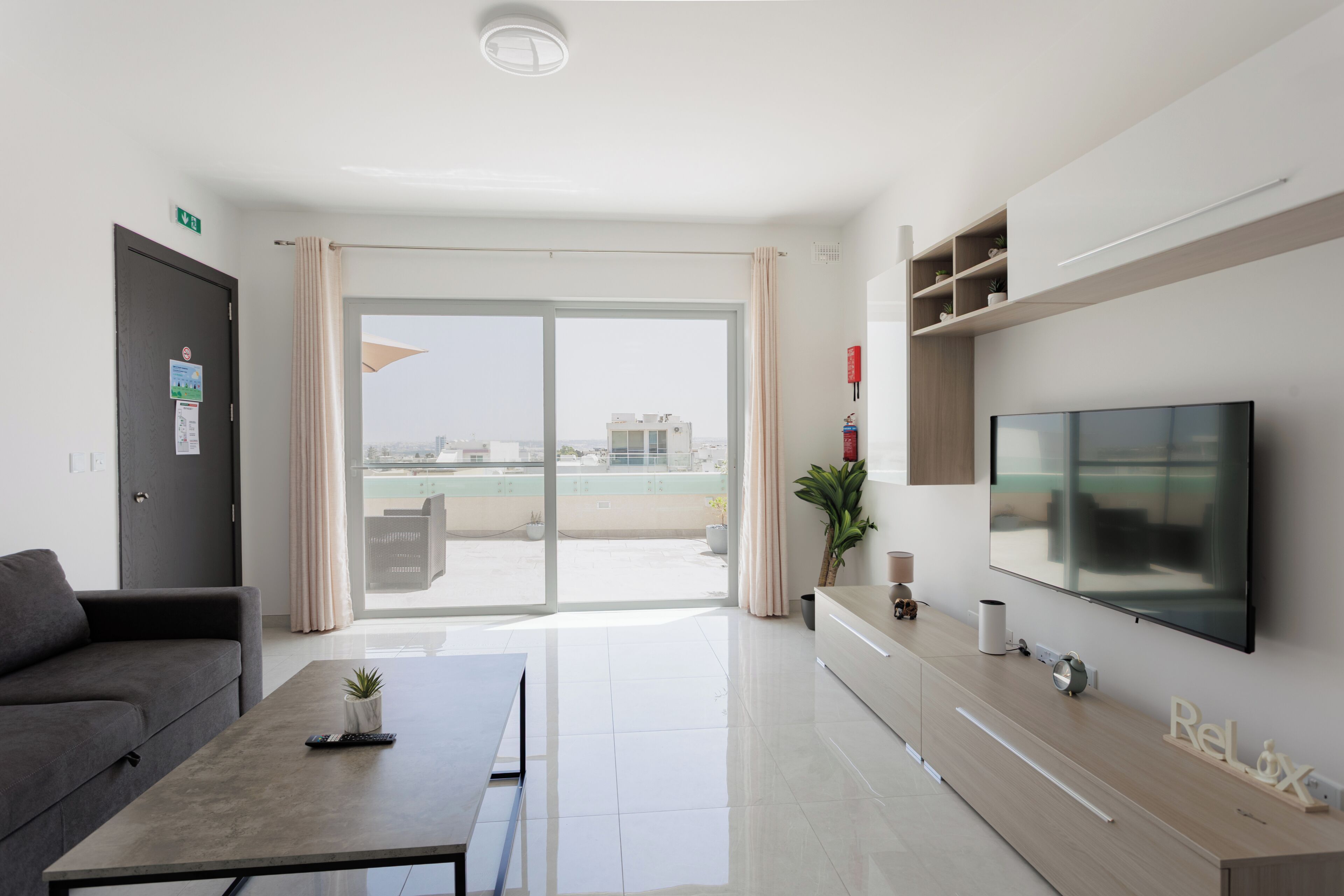 Habitación de hotel moderna con un televisor de pantalla plana grande, un sofá, una mesa de centro y un ventanal corredero que da a un balcón.