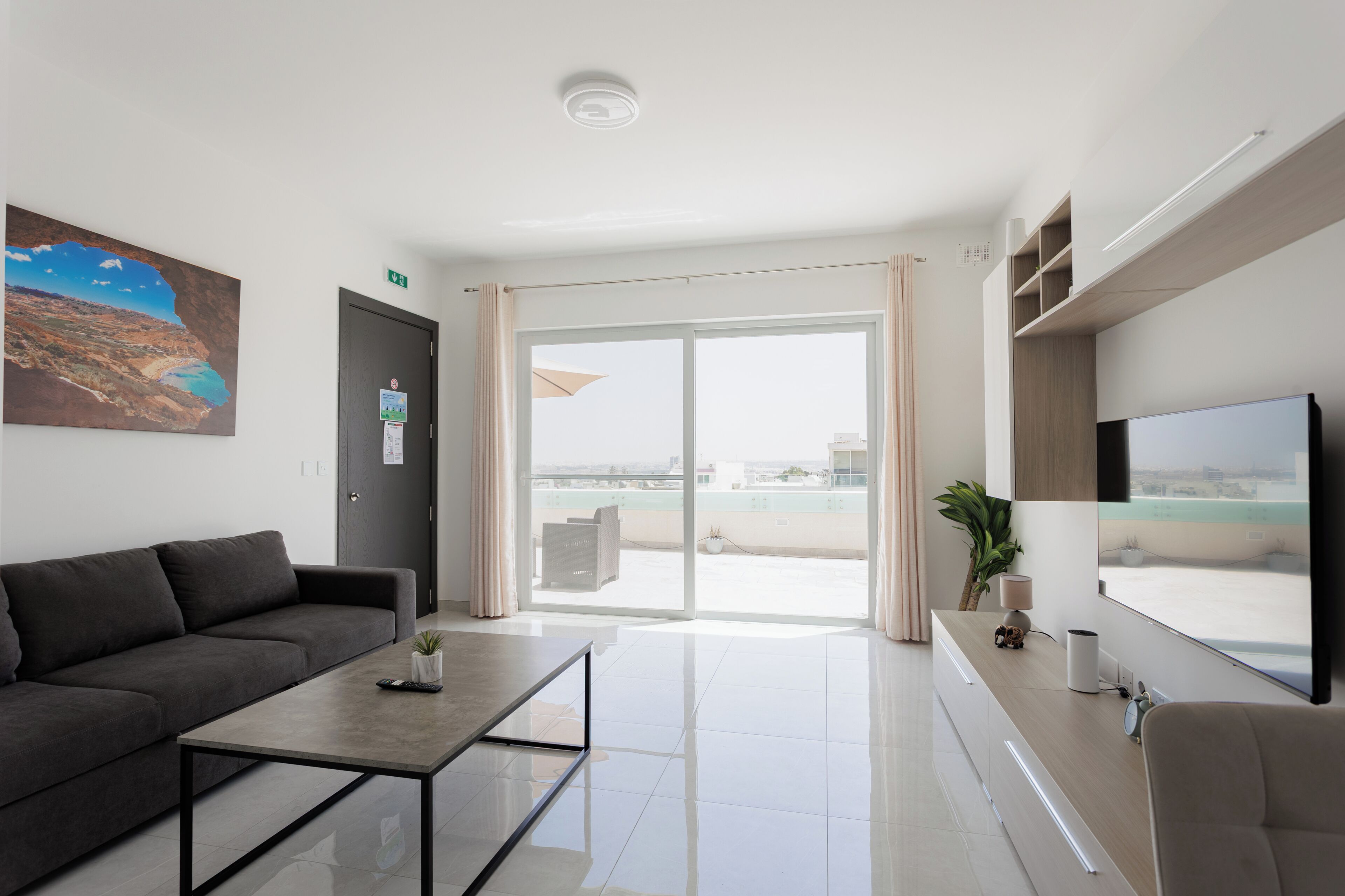 Una habitación de hotel moderna con sofá, una mesa de centro, televisor, balcón con vista a la ciudad y un cuadro en la pared.