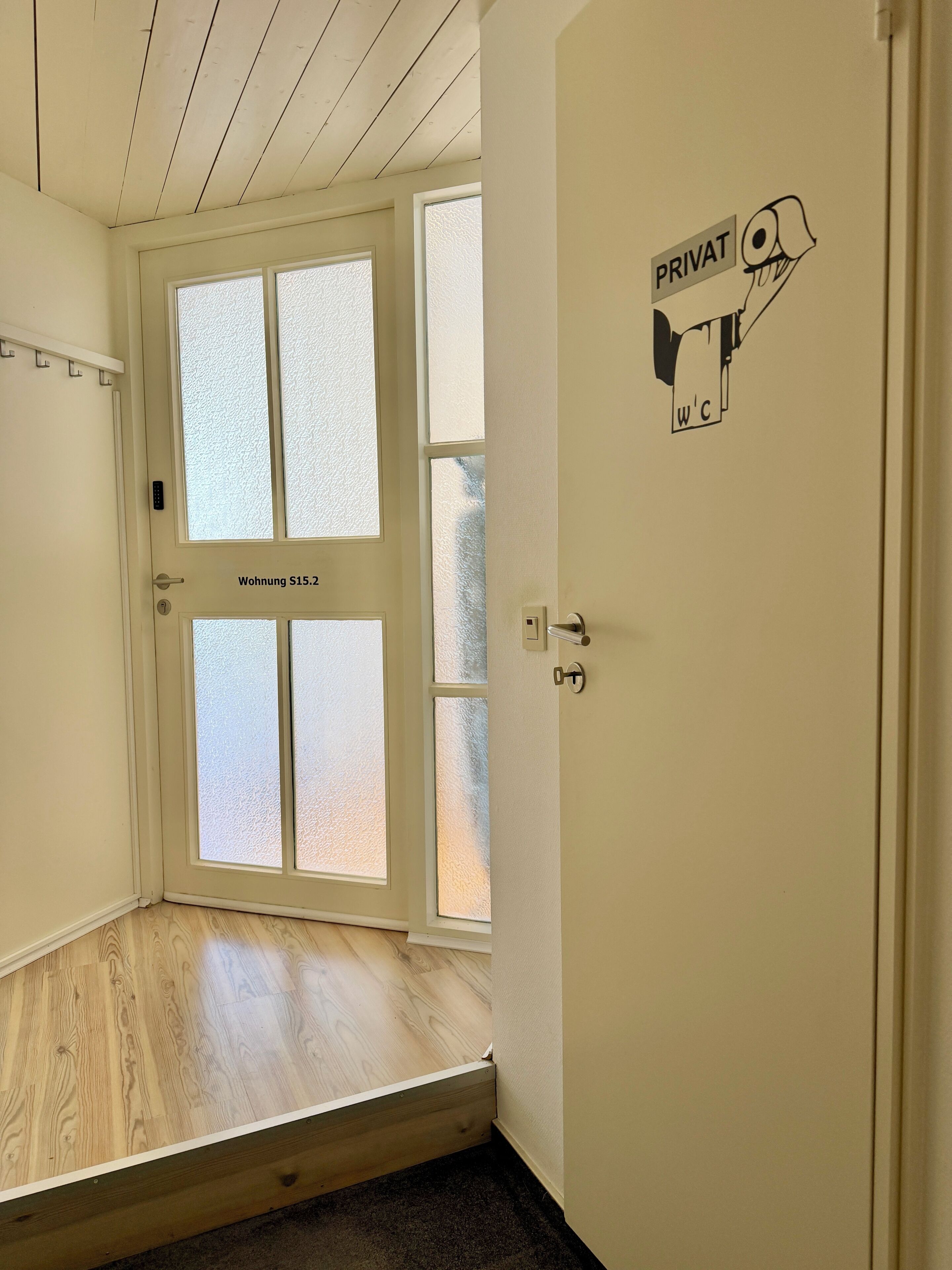 Een kamer met een witte deur waarop 'Wohnung S15.2' staat, geëtste glazen ramen, een houten vloer en een bordje 'PRIVAT WC'.