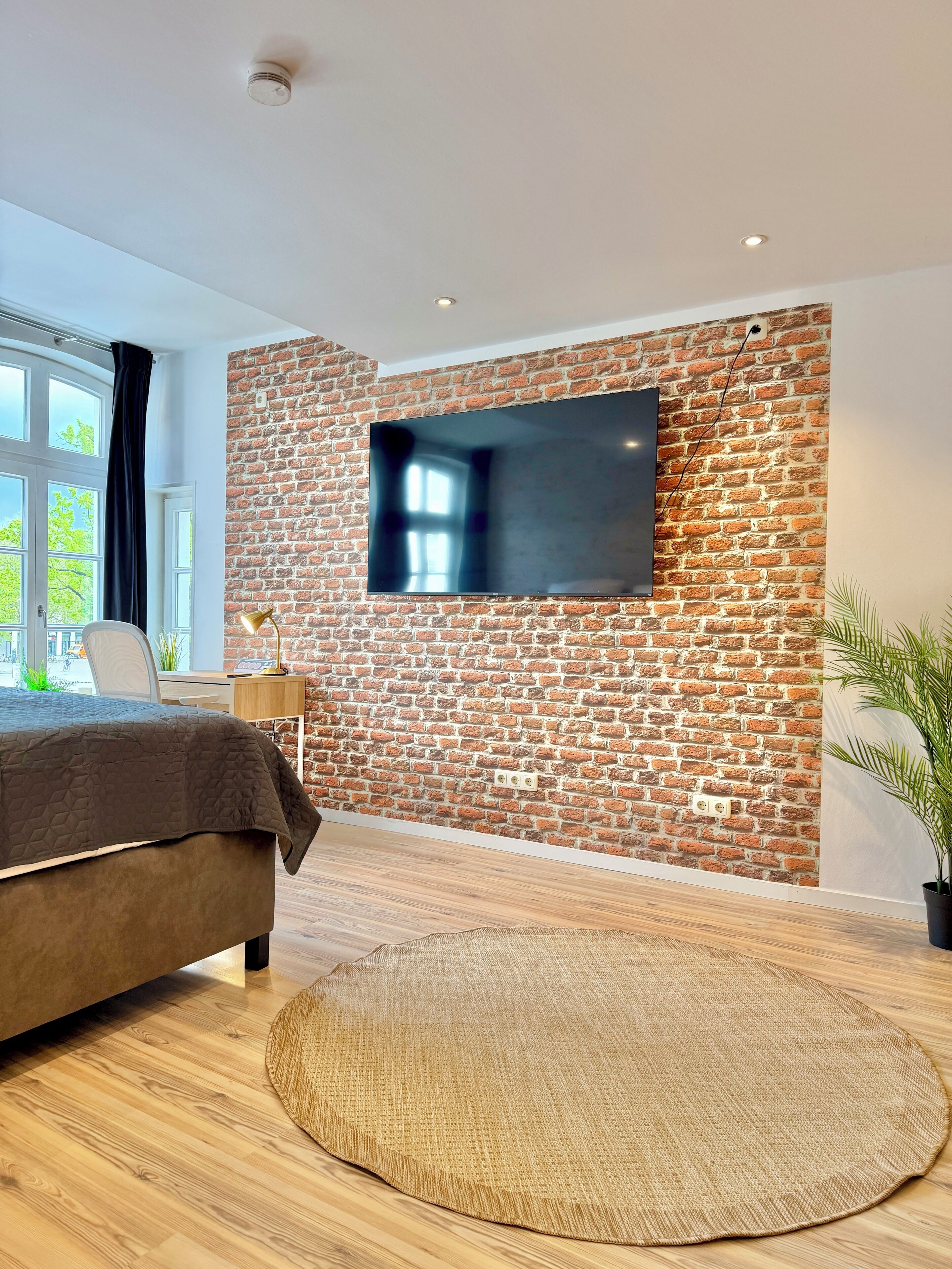 Une chambre avec un mur en briques, un téléviseur à écran plat, un lit, un tapis rond tissé et un bureau avec une chaise.