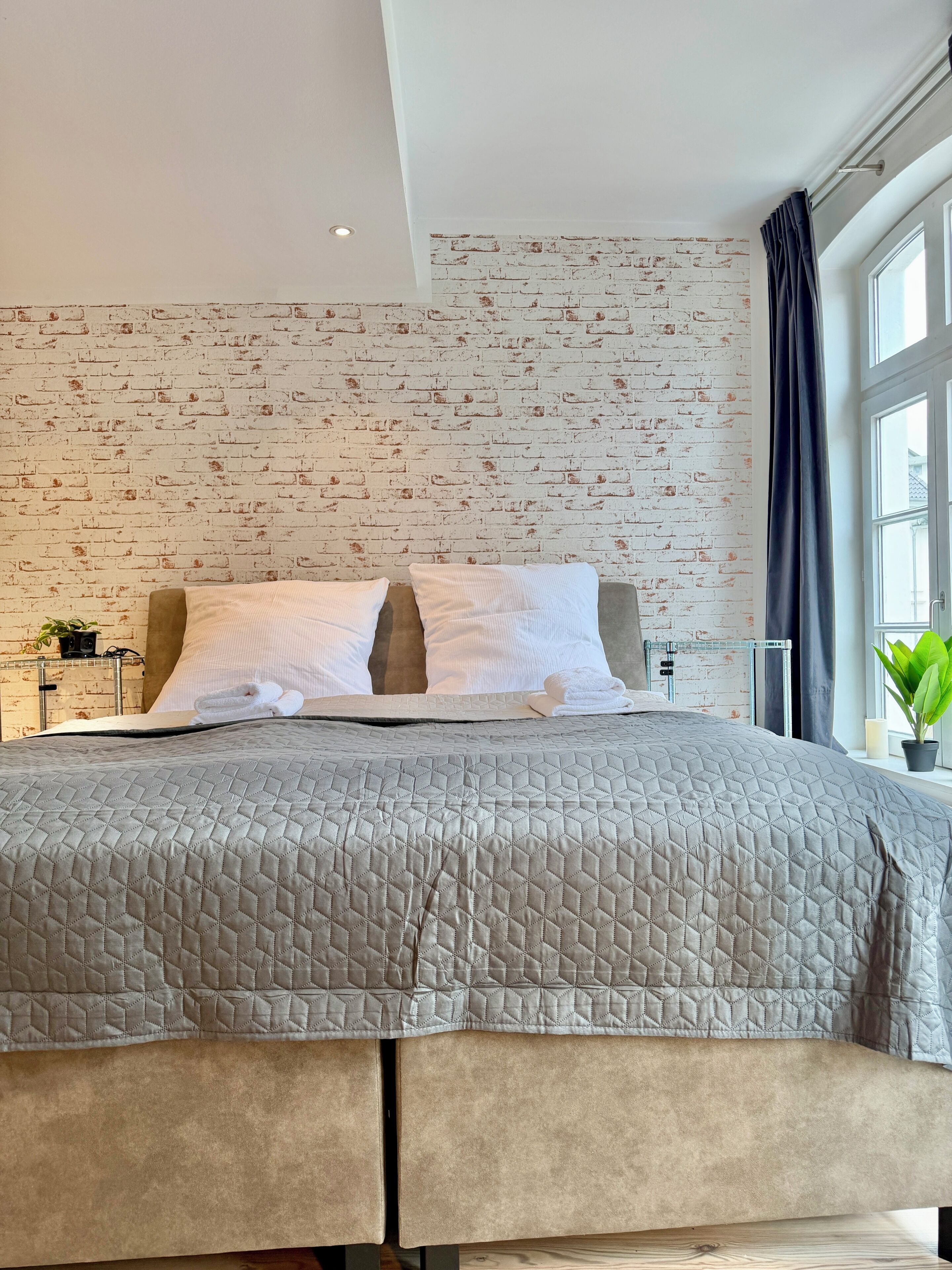 Un dormitorio moderno con una pared de ladrillo, una cama grande con cubrecama acolchado y una ventana con cortina.