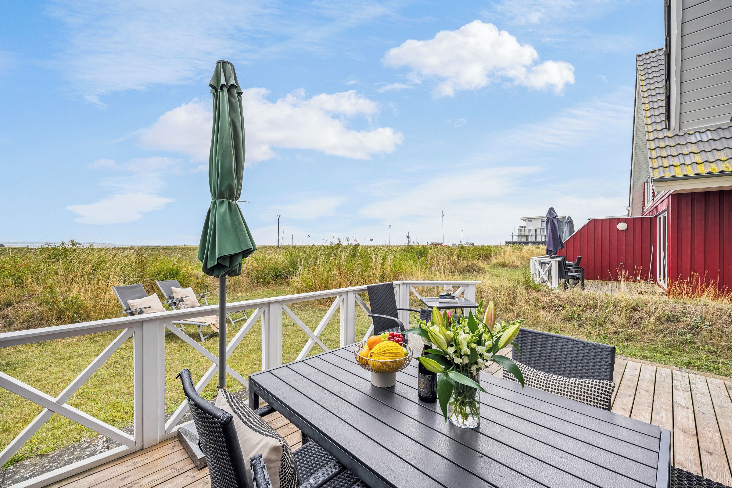 Una terraza con mobiliario exterior, una mesa con fruteros y flores, y un edificio rojo al fondo.