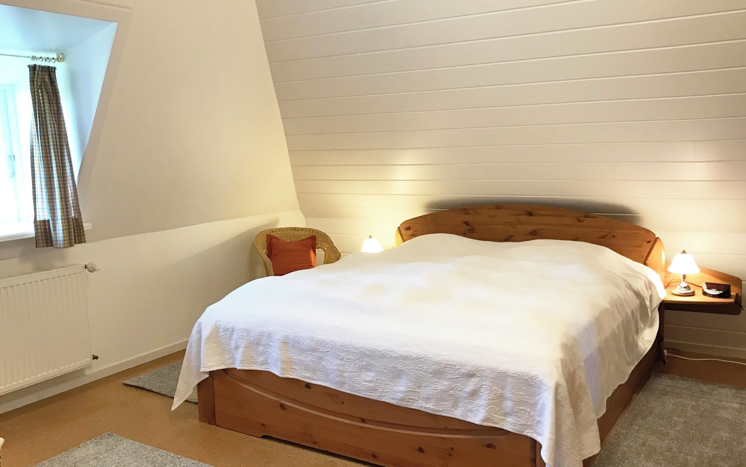 Ein Schlafzimmer mit einem hölzernen Bett, einem Nachttisch mit Lampe, einem Fenster mit Vorhängen und einer Heizung.