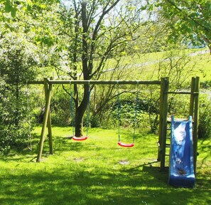 Un terrain de jeux avec des balançoires et un toboggan, entouré d’herbe verte et d’arbres.