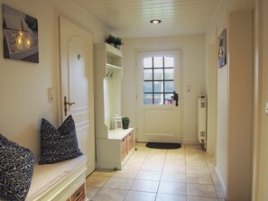 A hallway with a white door, a bench with cushions, a small table with a plant, and a window with a view of buildings outside.