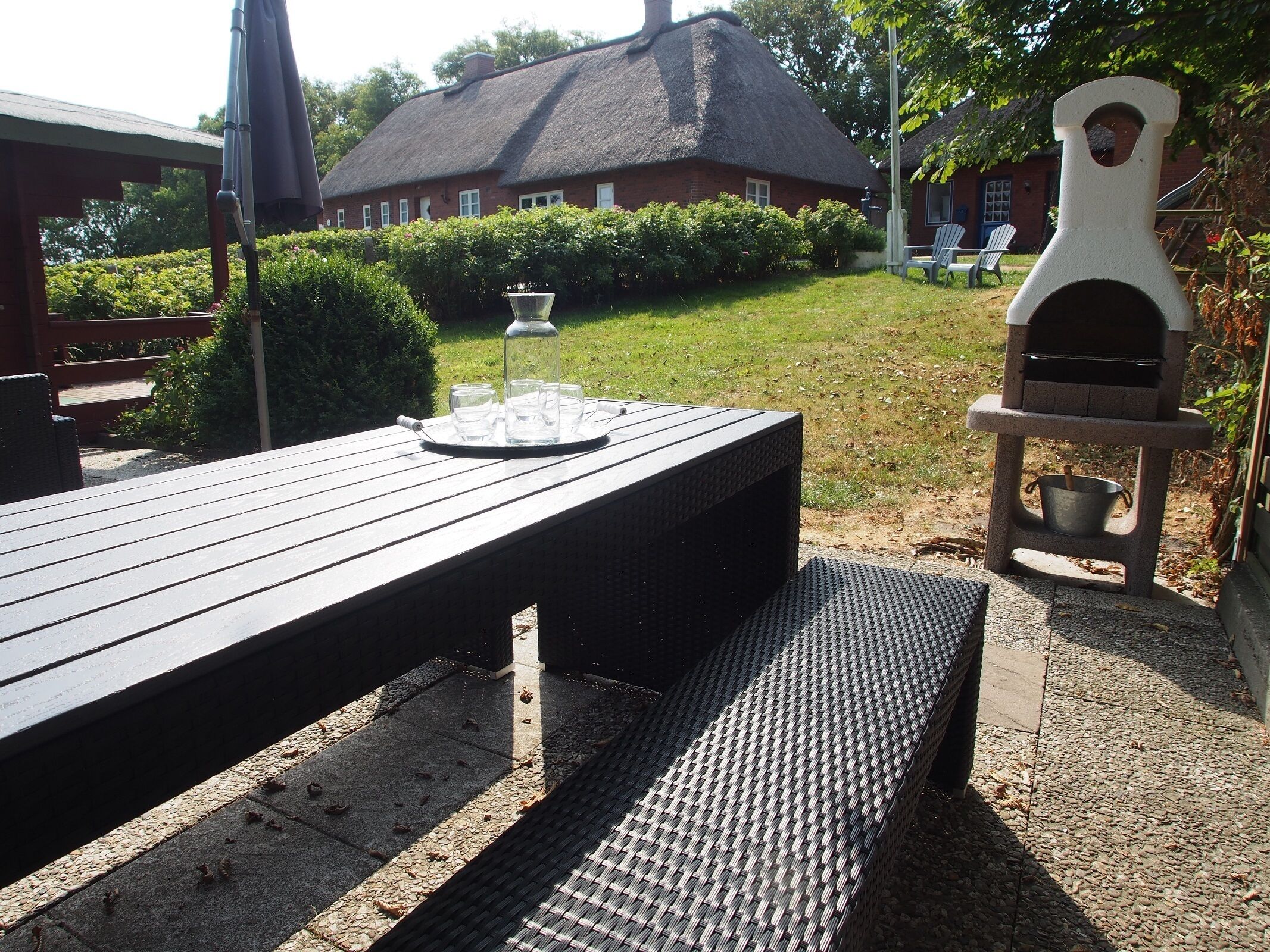 Eine Terrasse im Freien mit einem Holztisch, einem Grill und einem Gebäude mit einem Reetdach im Hintergrund.