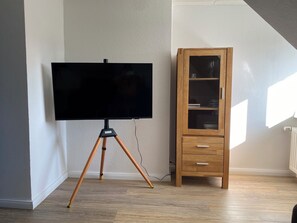 Ein Zimmer mit Fernseher auf einem Stativ, einem Holzkasten mit Schubladen und einem Heizkörper an der Wand.
