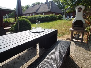 An outdoor patio with a wooden table, a grill, and a thatched-roof building in the background. - Wattwurm – shared Washing machine, use of garden, 2 floors, barbecue, parking (Ockholm)