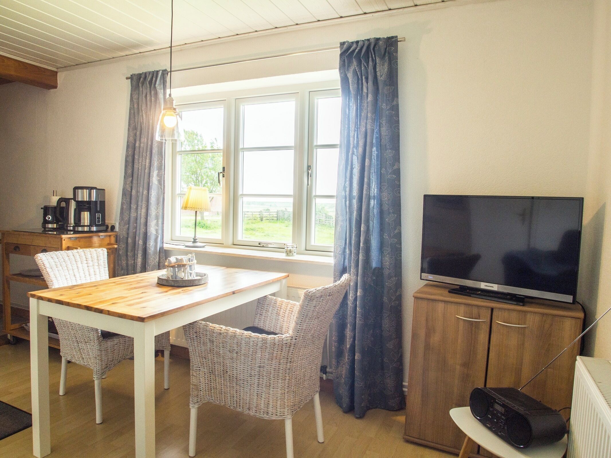 Une pièce comprenant une table en bois, des chaises en osier, un téléviseur, une machine à café et une radio.