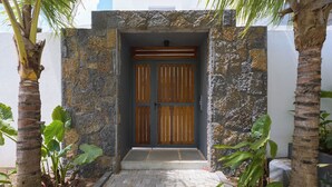 Uma entrada de pedra com porta de madeira, palmeiras e um caminho pavimentado que leva a um edifício.