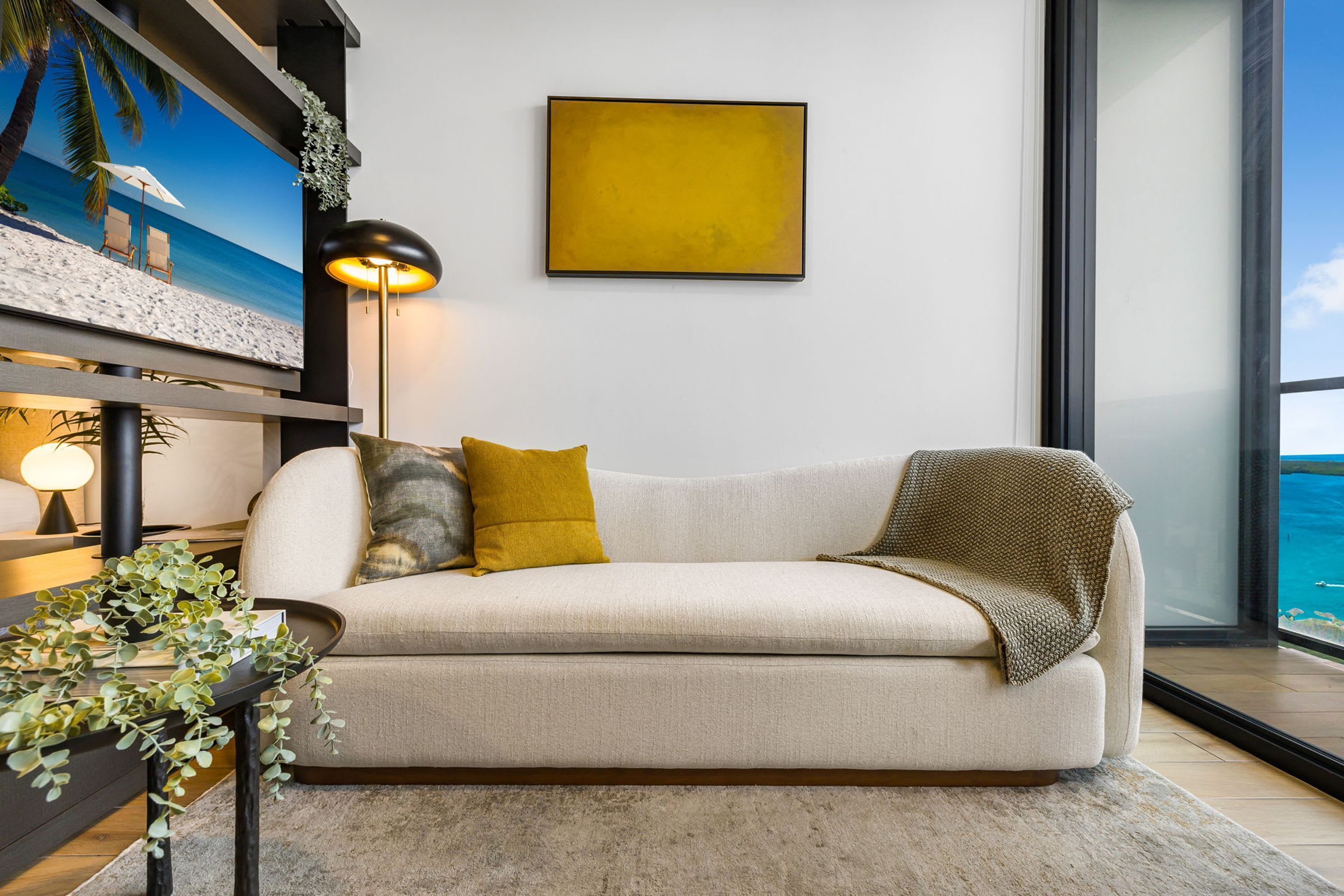 A modern living room with a sofa, a painting, a lamp, and a view of the ocean.