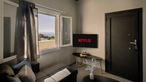 A modern living room with a sofa, a TV displaying 'NETFLIX,' a door, and a view of the outdoors. - Sunset Studio Seaview - Nea Premium Residences (Santorini)