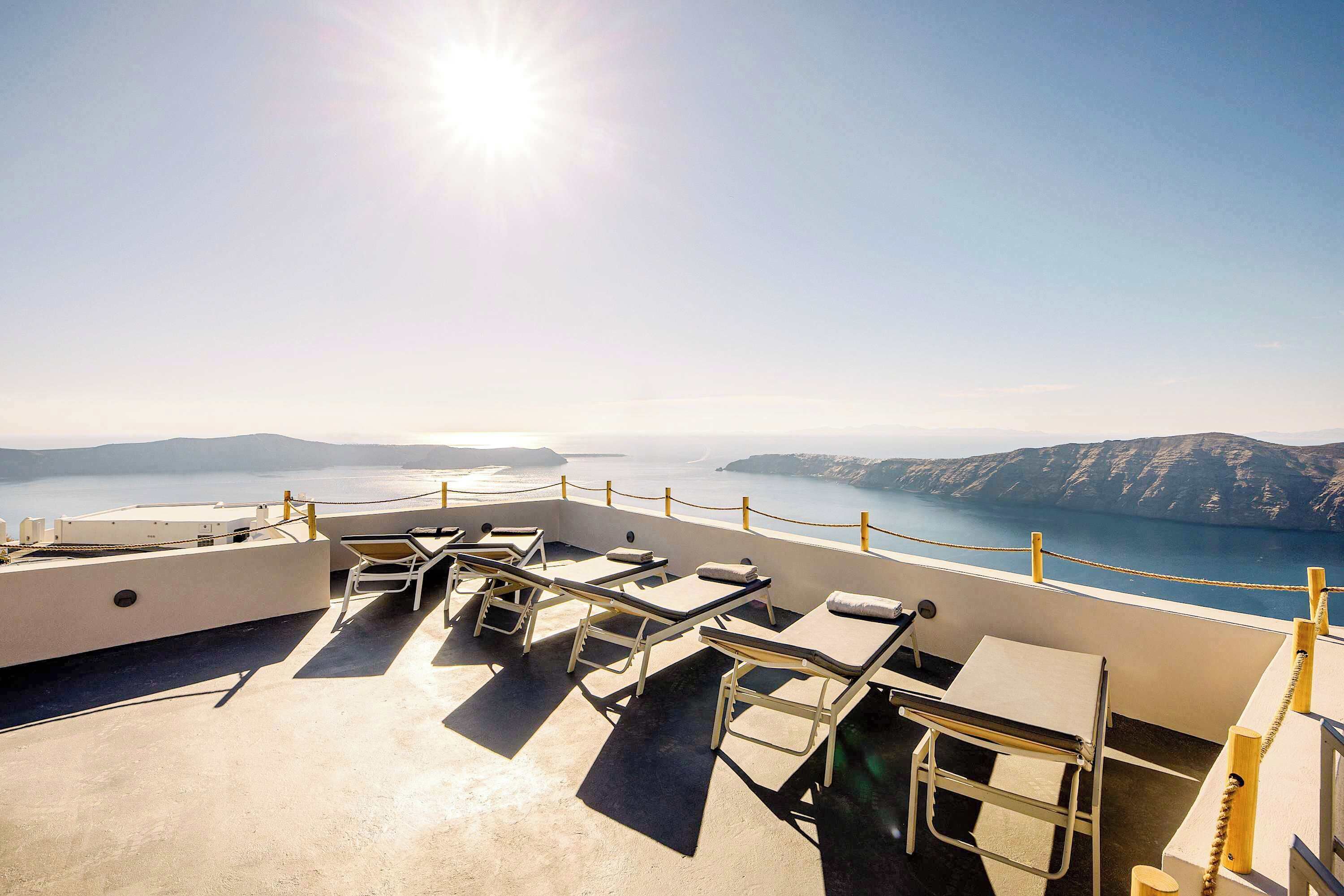A rooftop terrace with sun loungers, a table, and a view of the sea and mountains.