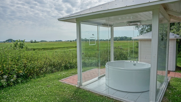Une salle de bain extérieure entièrement vitrée, dotée d’un jacuzzi, entourée d’une pelouse et de champs.