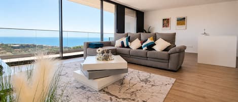 A modern living room with a large window, a grey sofa, a white coffee table, and a view of the sea.