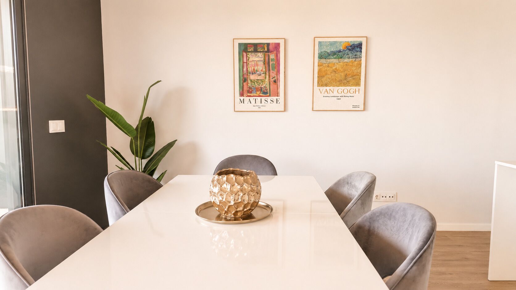 Ein Essbereich mit einem weißen Tisch, vier Stühlen und einer dekorativen goldfarbenen Schale. Zwei gerahmte Kunstwerke hängen an der Wand.