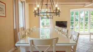 Uma sala de jantar com mesa de vidro, cadeiras brancas, um lustre, televisão e janelas amplas.